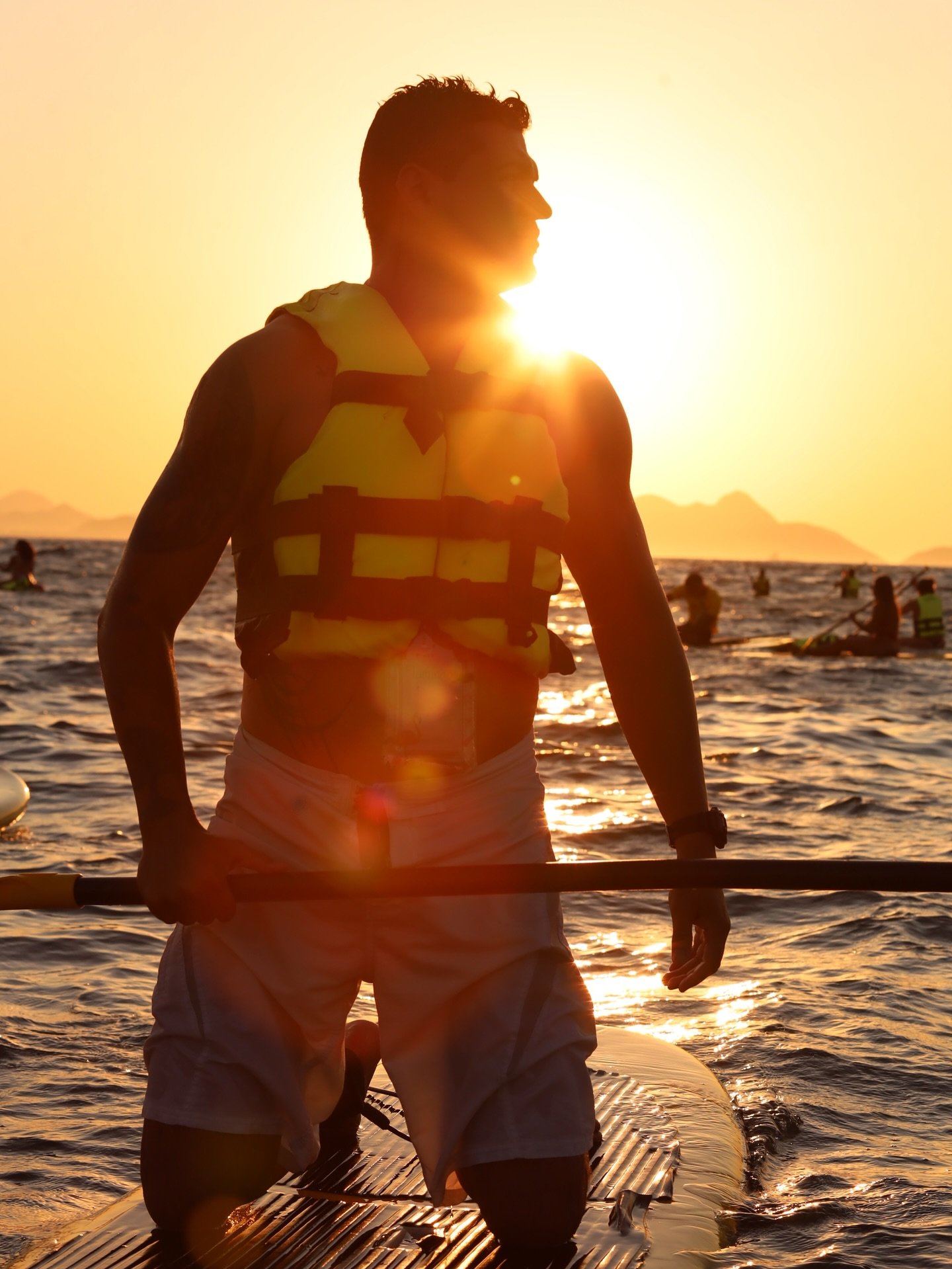 Homem posando de joelhos na prancha de stand up paddle em Copacabana com o nascer do sol e outras pessoas remando ao fundo.