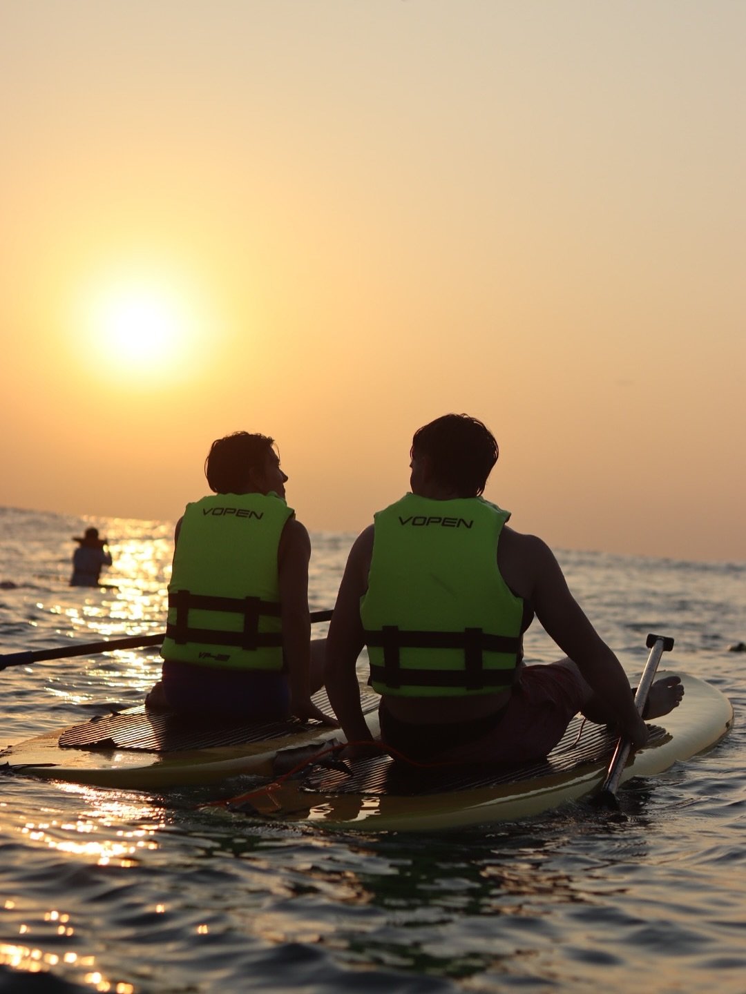 Casal de costas, cada um em sua prancha de stand up paddle em Copacabana, com coletes e o nascer do sol ao fundo.