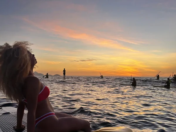 Mulher sentada na prancha de SUP olhando para o horizonte durante um nascer do sol laranja e roxo no Rio de Janeiro