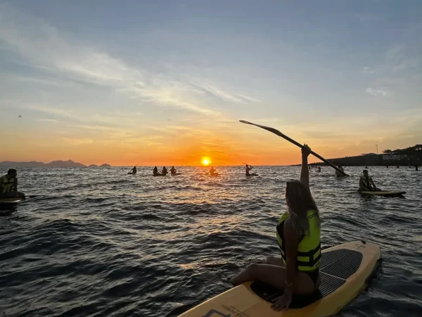 Mulher de costas na prancha de SUP levantando o remo em direção ao sol nascendo no horizonte em Copacabana